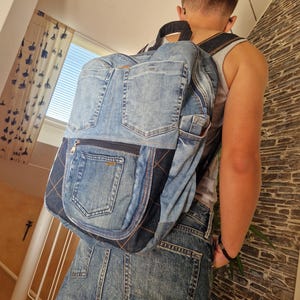 May include: A backpack constructed from repurposed blue denim fabric featuring multiple exterior pockets, including two prominent back pockets from jeans, dark blue denim panels with orange contrast stitching, and adjustable shoulder straps.