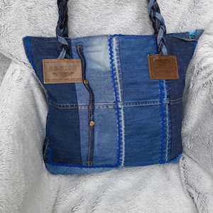 Könnte beinhalten: Eine Tragetasche aus Patchwork-Denim in verschiedenen Blautönen mit geflochtenen Denim-Henkeln, blauen Ziernähten und zwei braunen lederartigen Aufnähern. Ein Aufnäher ist mit dem Text AS-KISS geprägt.
