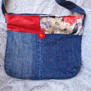 Könnte beinhalten: Eine Umhängetasche aus blauem Denim mit einem rautenförmigen Steppmuster aus rotem Garn. Der obere Bereich besteht aus einem einfarbigen roten Stoffstreifen und einem floralen Muster in Rot, Gelb und Grau, verziert mit einem runden roten Knopf.