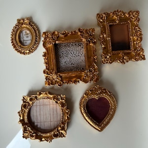 May include: A collection of ornate, gold-colored miniature picture frames in various shapes. The frames include oval, square, and heart shapes, with decorative scrollwork and textured backgrounds. Some frames have patterned inserts.