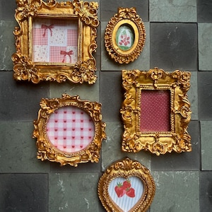 May include: A collection of ornate, gold-colored picture frames in various shapes, including square, oval, and heart. The frames hold images with patterns like gingham, polka dots, and strawberries. The frames are arranged on a textured, dark gray background.