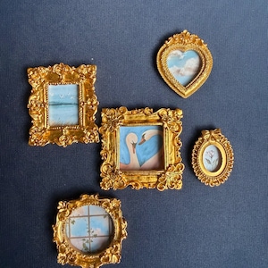 May include: A collection of miniature gold-framed art pieces. The frames are ornate and feature various paintings, including a heart-shaped frame with a sky scene, and a square frame with two swans. The art pieces are arranged on a dark blue surface.