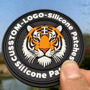 May include: A circular black silicone patch with a tiger's face in orange, white, and black. The patch has the text "CUSTOM-LOGO-Silicone Patches" in white around the edge. The patch is held by a hand.
