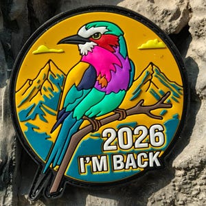Op de afbeelding: Een ronde patch met een kleurrijke vogel op een tak, tegen een gele achtergrond met bergen en wolken. De tekst "2026 I'M BACK" staat in het wit. De patch heeft een zwarte rand.