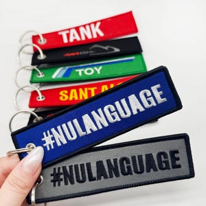 Puede incluir: Varios llaveros bordados en colores variados, incluyendo rojo, negro, verde y azul. Cada llavero presenta texto blanco, con el llavero azul mostrando "#NULANGUAGE". Los llaveros están unidos a anillos metálicos plateados.