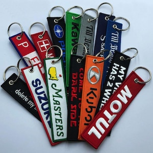 May include: A collection of embroidered keychains in various colors, including red, black, green, and orange. Each keychain features text or logos, such as "MOTUL", "Masters", and "Suzuki", with a metal ring for attachment. The keychains are arranged in a fan shape.