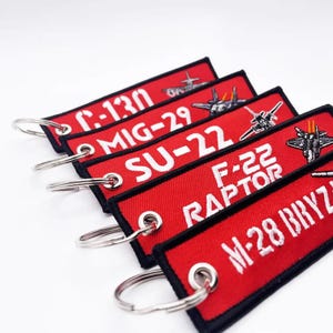 May include: Red embroidered keychains with white text and silver keyrings. The keychains feature the names of military aircraft: C-130, MIG-29, SU-22, F-22 RAPTOR, and M-28 BRYZ. Each keychain has a black border and a small image of the aircraft.