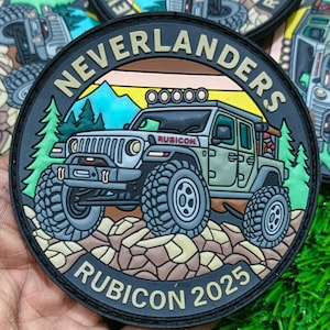 May include: A circular patch with the words "NEVERLANDERS" and "RUBICON 2025". The design shows a green off-road vehicle on rocks, with a mountain and forest backdrop. The patch is black with a detailed design.