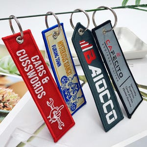 May include: Four embroidered keychains with silver rings. The keychains are red, blue, green, and gray. The red keychain says "CARS & CUSSWORDS" with a wrench graphic. The blue keychain says "MYSTERIOUS XIANGKI FURONG TOWN". The green keychain says "AIOCCO". The gray keychain says "CAFECITO espresso coffee".