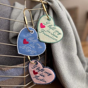 May include: Three heart-shaped keychains with embroidered text and a small heart design. The keychains are in blue, pink, and green, with the phrases "I left my in NYC", "I left my in San Francisco", and "I left my in Austin". They hang from gold-colored clasps.