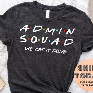 May include: Dark gray t-shirt with the words "ADMIN SQUAD" in a white, stylized font, with colored dots. Below, the text reads "WE GET IT DONE". The shirt has a crew neck and short sleeves.
