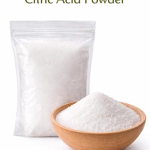 以下が含まれることがあります： 白いクエン酸パウダーが入った透明なビニール袋と木製のボウル。画像上部には緑色で「Citric Acid Powder」の文字が表示されています。パウダーは粒状で、食品成分のようです。