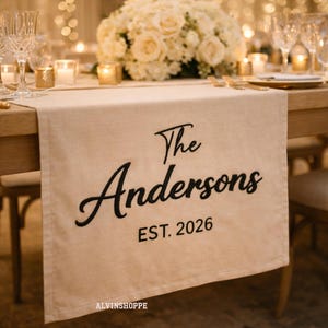 Puede incluir: Camino de mesa beige con texto negro "The Andersons EST. 2026". El camino de mesa está sobre una mesa de madera para un evento formal, con un arreglo floral y velas.