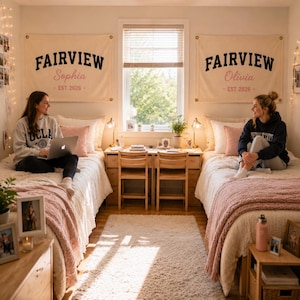Op de afbeelding: Twee crèmekleurige stoffen wandbanners met de tekst FAIRVIEW in het zwart, de namen Sophia of Olivia in roze cursief schrift, en de tekst EST 2026 eronder. De banners hangen boven twee eenpersoonsbedden in een gedeelde slaapkamer.