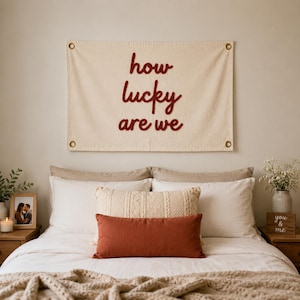 Puede incluir: Un tapiz de pared color crema con la frase "how lucky are we" en escritura roja. La pancarta está sobre una cama con ropa de cama blanca, almohadas decorativas y una manta tejida. Una foto enmarcada y flores están en las mesitas de noche.
