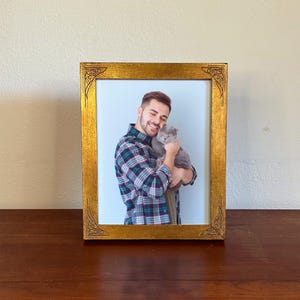 May include: A rectangular picture frame with a metallic gold finish and decorative corner detailing. The frame contains a photograph of a person with a beard wearing a plaid shirt, holding a grey cat with orange eyes against a light background.
