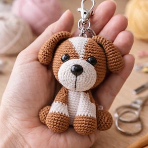 May include: A handmade crochet dog keychain in brown and white. The small stuffed animal has a white face and chest, brown ears and paws, and black eyes. It is attached to a silver-colored metal clasp.