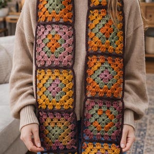 May include: A colorful, crocheted scarf featuring a granny square design. The scarf showcases squares in shades of orange, green, pink, and yellow, with a dark brown border. The scarf is a handmade accessory.