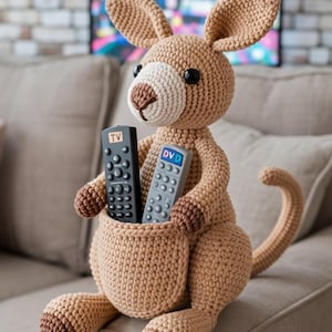 Pode incluir: Um brinquedo de crochê bege feito à mão em forma de canguru, com um bolso contendo dois controles remotos. Um diz "TV" e o outro "DVD". O canguru tem orelhas grandes, um focinho branco e olhos escuros. O brinquedo está em um sofá.