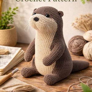 以下が含まれることがあります： 茶色とベージュの色合いの、かぎ針編みのカワウソのぬいぐるみ。木製の表面に置かれています。「Otter Crochet Pattern」の文字がぬいぐるみの上に表示されています。カワウソは、黒い鼻とひげを持つ詳細なデザインです。