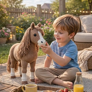 Op de afbeelding: Een gehaakt bruin paard met een witte kop en manen staat in de buurt van een kind. Het kind zit op een kleed en reikt naar het paard. Een houten dienblad bevat een gele mok, koekjes, aardbeien en een fles sap.