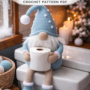 Könnte beinhalten: Ein gehäkelter Zwerg mit blauem Hut und Bart, der eine Rolle Toilettenpapier hält. Der Zwerg sitzt auf einer weißen Oberfläche. Der Text "Winter Bathroom Gnome Crochet Pattern PDF" befindet sich oben im Bild.