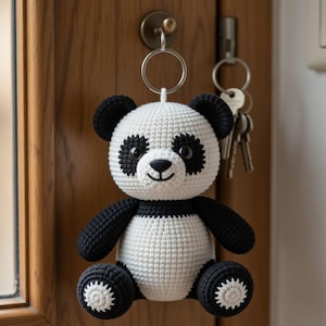 May include: A handmade crochet panda keychain. The panda has a white body with black accents on the ears, arms, legs, and around the eyes. The keychain is hanging on a door with keys.