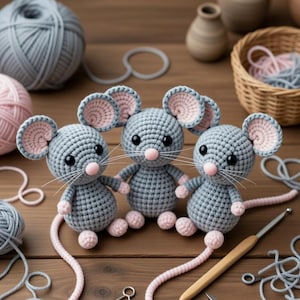 Puede incluir: Tres ratones de ganchillo hechos a mano en gris con detalles rosas, incluyendo orejas, patas y colas. Los ratones están sentados sobre una superficie de madera, con hilo, un ganchillo y otros suministros de artesanía. Un artículo encantador, hecho a mano.