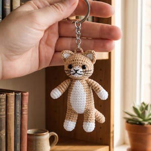 May include: A crocheted cat keychain in tan and white. The handmade cat has a smiling face with black whiskers and eyes. The keychain is held up in front of a bookshelf with books and a mug.