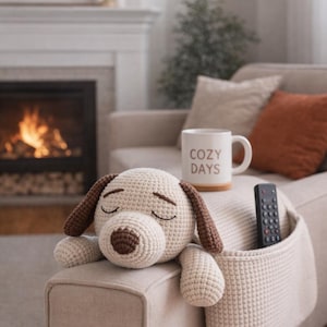 Pode incluir: Um cão de pelúcia de crochê com orelhas e focinho marrons descansa em um sofá bege. Uma caneca branca com "COZY DAYS" impressa está por perto. Um controle remoto está em um bolso de crochê no sofá.