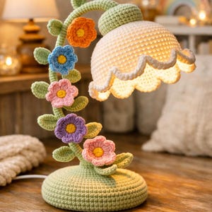 Puede incluir: Una lámpara de ganchillo hecha a mano con un diseño floral. La lámpara presenta una base y un tallo de color verde claro, adornados con flores de ganchillo coloridas en naranja, azul, rosa y morado. La pantalla es blanca con un borde festoneado.