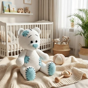May include: A handmade white crochet teddy bear with blue accents on its paws, ears, and nose. The bear is sitting on a cream-colored blanket, with a wooden rattle and a beige ball nearby. The scene suggests a nursery or children's room.
