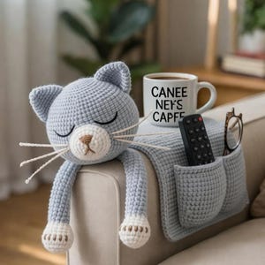 May include: A light blue crocheted cat-shaped sofa armrest organizer with a pocket for a remote control and glasses. The cat has closed eyes, white whiskers, and white paws. A mug with the text "CANEE NE'S CAPFE" sits on the armrest.