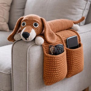 May include: A crocheted dog-shaped couch organizer in shades of brown, white, and black. The dog has long ears, a white chest, and a tail. Two pockets hang down, holding a remote, glasses, and a phone. The organizer rests on a gray couch.