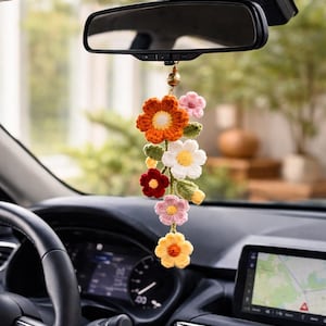 May include: A colorful, handmade crochet car charm featuring a cascade of flowers in shades of orange, pink, white, yellow, and red. The charm hangs from a gold-colored bell and is suspended from a car's rearview mirror.