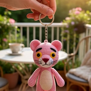 May include: A pink crocheted panther keychain with black accents and yellow eyes is held up by a hand. The keychain is attached to a silver chain and ring. The background includes a table, chairs, and greenery.