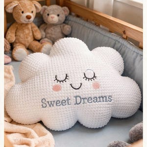 Könnte beinhalten: Weißes wolkenförmiges Kissen mit gestickten geschlossenen Augen, rosigen Wangen und dem Text "Sweet Dreams" in Hellblau. Zwei Teddybären sitzen im Hintergrund. Das Kissen liegt in einem Kinderbett.