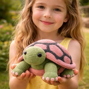 Puede incluir: Una tortuga de ganchillo con un caparazón rosa y gris, cabeza y extremidades verdes y ojos negros. La tortuga es sostenida por una persona que lleva un vestido amarillo. El fondo es verde borroso.