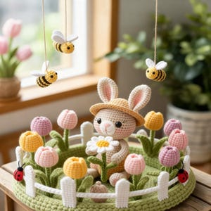 Op de afbeelding: Een gehaakte paashaas met een hoed, die een madeliefje vasthoudt, omringd door gehaakte tulpen in een groene tuin. Inclusief hangende gehaakte bijen en een wit hek. De tulpen zijn in tinten geel, roze en paars.