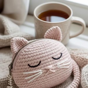 Puede incluir: Un monedero con forma de gato de ganchillo rosa claro con cierre de cremallera, ojos bordados negros y bigotes blancos. El monedero tiene un pequeño dije de espina de pescado en el tirador de la cremallera. Una taza de té está en el fondo.