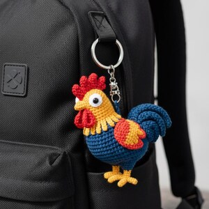Può includere: Un portachiavi a forma di gallo all'uncinetto dai colori vivaci è appeso a uno zaino nero. Il gallo ha una cresta rossa, testa gialla, corpo blu e dettagli arancioni e gialli. Il portachiavi è attaccato a un portachiavi argentato.