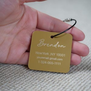 Könnte beinhalten: Ein goldfarbener quadratischer Anhänger mit dem Namen "Brendon" in Schreibschrift. Darunter stehen die Worte "New York, NY 10001", eine E-Mail-Adresse und eine Telefonnummer. Der Anhänger ist an einer schwarzen Schnur befestigt.