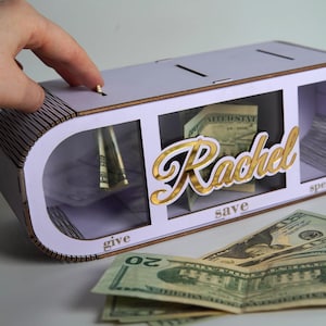 Puede incluir: Una hucha de madera de color lila claro con tres compartimentos etiquetados como "give", "save" y "spend". La palabra "Rachel" está en letras doradas. Varios billetes de dólar estadounidense son visibles dentro y fuera.