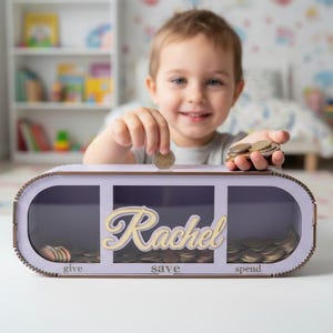 Può includere: Un salvadanaio in legno color lavanda con tre scomparti: "give", "save" e "spend". Il nome "Rachel" è scritto in corsivo chiaro. Un bambino sta mettendo delle monete nello scomparto "save".
