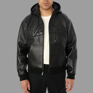 K&ouml;nnte beinhalten: Schwarze Lederjacke mit Kapuze und Rei&szlig;verschluss, mit gepr&auml;gtem Schriftzug auf der Brust. Die Jacke hat einen gerippten Saum und B&uuml;ndchen sowie eine Vordertasche. Das Model tr&auml;gt ein wei&szlig;es T-Shirt darunter.