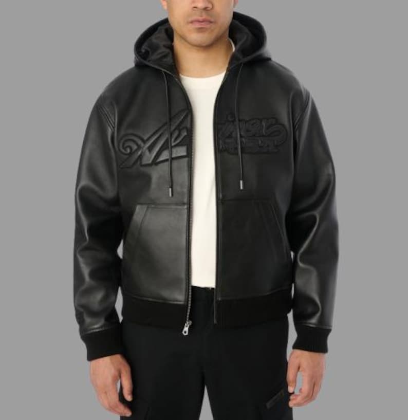 K&ouml;nnte beinhalten: Schwarze Lederjacke mit Kapuze und Rei&szlig;verschluss, mit gepr&auml;gtem Schriftzug auf der Brust. Die Jacke hat einen gerippten Saum und B&uuml;ndchen sowie eine Vordertasche. Das Model tr&auml;gt ein wei&szlig;es T-Shirt darunter.