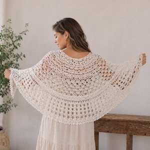 May include: A cream-coloured, open-weave crochet shawl with a scalloped edge. The shawl is draped over the shoulders, showcasing its intricate design and airy texture. The shawl's delicate pattern and neutral colour make it a versatile accessory.