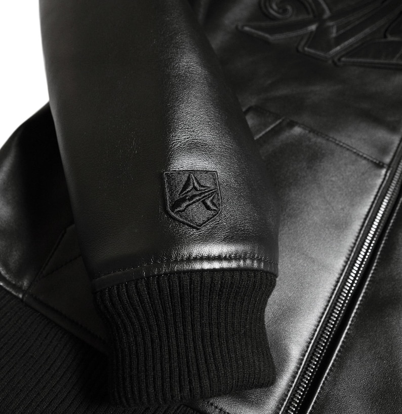 K&ouml;nnte beinhalten: Nahaufnahme einer schwarzen Lederjacke. Die Jacke hat eine gerippte Manschette, ein kleines Emblem am &Auml;rmel und einen Rei&szlig;verschluss. Das Leder hat einen leichten Glanz, und die N&auml;hte sind sichtbar. Die Jacke scheint neu zu sein.