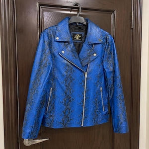 May include: A bright blue motorcycle jacket featuring a black snakeskin pattern, a notched collar, silver-tone metal zippers on the front and pockets, and snap button details. The interior label displays the text 'Vera Couture' in gold lettering.