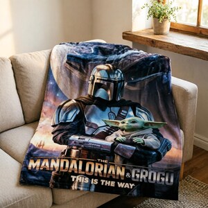 May include: A soft blanket depicting a scene from The Mandalorian series. The design shows the Mandalorian character with Grogu, and the text "THE MANDALORIAN & GROGU THIS IS THE WAY". The blanket is draped over a beige sofa.